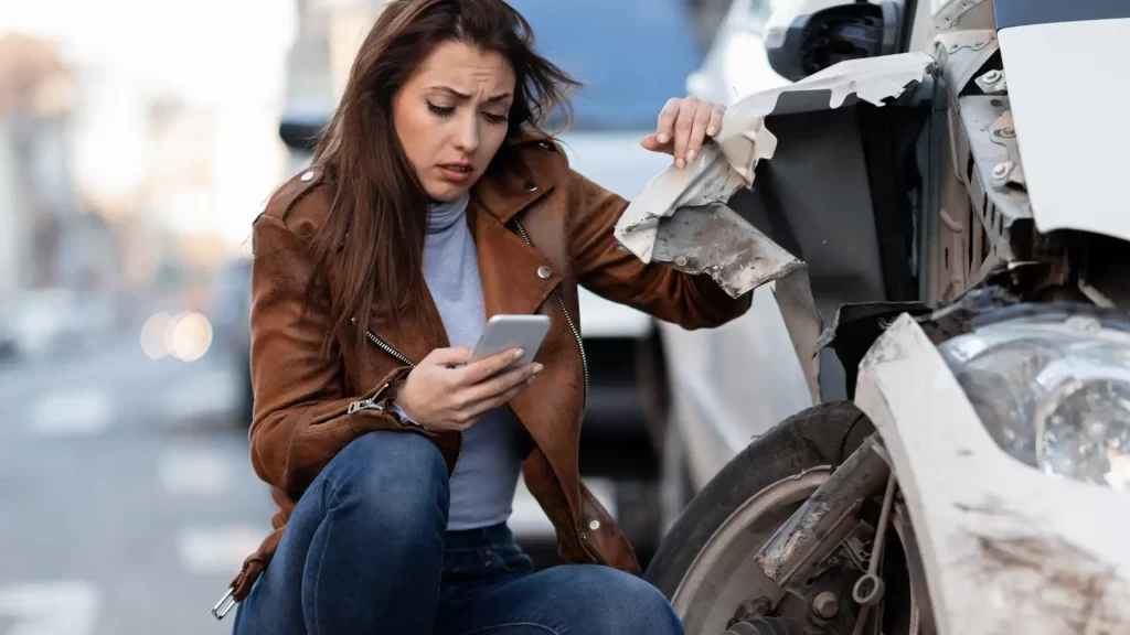 a woman looking at a phone next to a damaged hit and run car